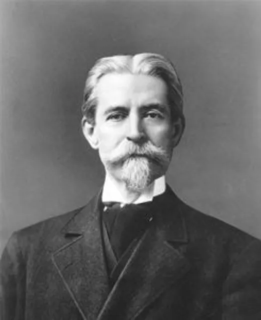 Black and white photograph of J.E. Hanger. He is a middle aged man dressed in a black suit. His hair appears white or gray and is parted down the middle. He has a moustache and goatee. This image is from the early 1900s.